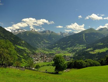 Ein üppig grünes Tal mit einer kleinen Stadt, umgeben von Bergen und einem teilweise bewölkten blauen Himmel.