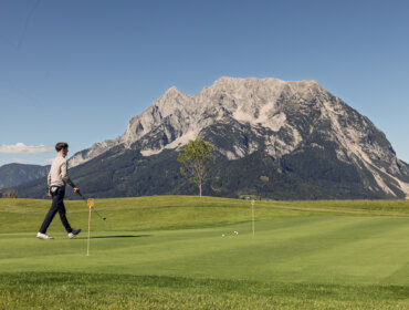 Christoph Haid (78) Ein Golfer spaziert auf dem grünen Platz des Golfclubs Schloss Pichlarn mit den Bergen im Hintergrund bei klarem Himmel.