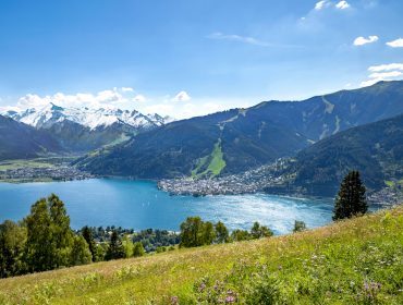 Blick auf einen See, umgeben von einer Stadt und Bergen mit schneebedeckten Gipfeln, blauer Himmel und grüne Wiese im Vordergrund.