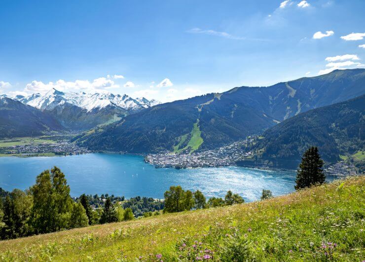 Blick auf einen See, umgeben von einer Stadt und Bergen mit schneebedeckten Gipfeln, blauer Himmel und grüne Wiese im Vordergrund.