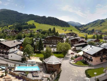 Ein malerisches Bergdorf mit Häusern im Chalet-Stil, grünen Hügeln und einem Schwimmbad des Wolf Hotels unter einem klaren Himmel.
