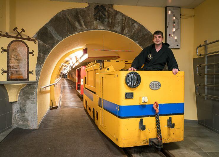 Ein Mann steht neben einer gelben Grubenlokomotive am Eingang eines Tunnels mit Bergbausymbolen darüber.