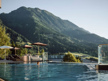An einem sonnigen Tag schwimmen Sie im Außenpool des Hotels Alpina St. Johann mit Bergen und Liegestühlen.