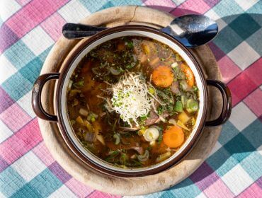 Schüssel mit Suppe mit Nudeln, Karotten und Kräutern auf einem Holzbrett, mit einem Löffel auf einer karierten Tischdecke.
