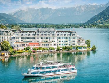 Ein weißes Boot fährt auf einem See in der Nähe des Grandhotels Zell am See, mit den Bergen im Hintergrund.