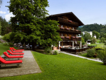 Hotel Zur Post Goldegg: Holzhotel mit Balkonen, roten Liegestühlen, Bäumen und grünen Hügeln unter blauem Himmel.