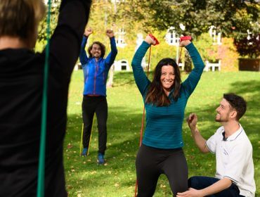 Vier Erwachsene trainieren mit Widerstandsbändern im Freien; eine Frau lächelt, während andere sie anfeuern und ihr helfen.
