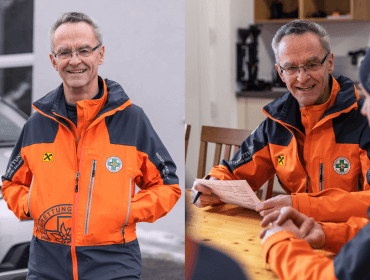 bergretter-balthasar-im-portrait Ein lächelnder Mann in einer orangefarbenen Rettungsjacke steht draußen und sitzt drinnen mit anderen an einem Tisch.