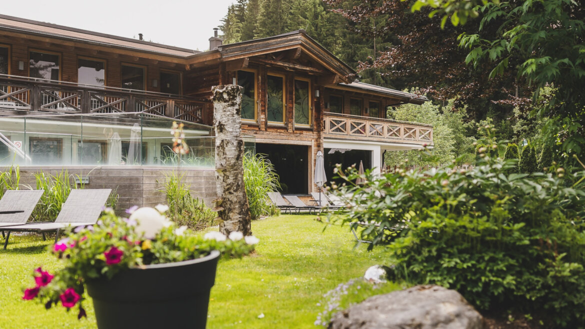 Die Holzhütte des Hotels Sonne mit ihren Balkonen liegt eingebettet zwischen grünem Gras, Bäumen und Gartenblumen in Töpfen. (vergrößerte Ansicht)
