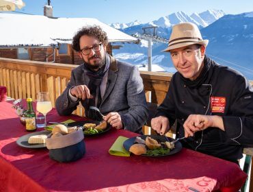 Zwei Männer beim Mittagessen an einem Tisch im Freien mit verschneiten Bergen im Hintergrund.