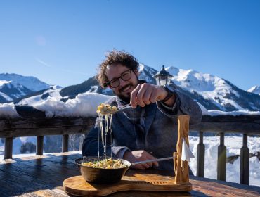 Rudi’s Roadtrip durch das SalzburgerLand – Winter Edition, Saalbach Ein Mann genießt Käsespätzle an einem verschneiten Bergtisch im Freien mit Blick auf schneebedeckte Berggipfel.