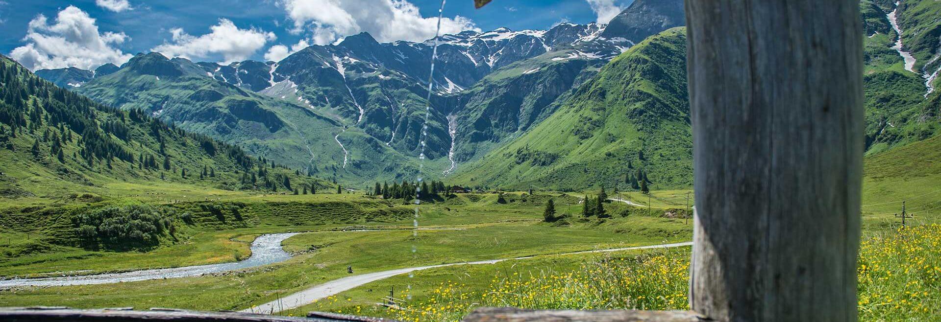 Ein hölzerner Brunnen lässt Wasser tropfen, während im Hintergrund Berge, ein grünes Tal und ein blauer Himmel zu sehen sind.