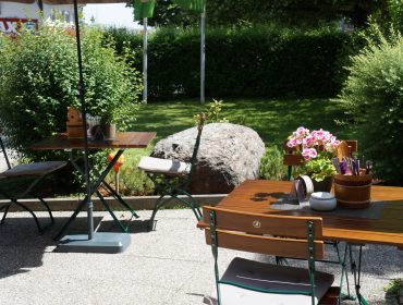 Tische und Stühle auf einer Terrasse im Freien, mit grünen Büschen und einem großen Felsen im Hintergrund.