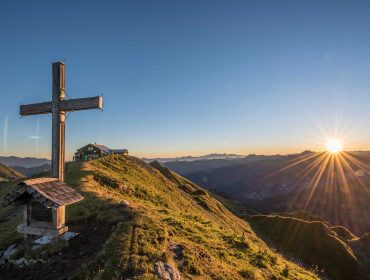 Großes Holzkreuz auf einem grasbewachsenen Berggipfel bei Sonnenaufgang, mit einer kleinen Schutzhütte und fernen Bergen.