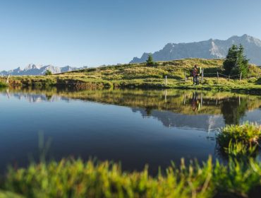 Grossarl Zwei Menschen stehen an einem ruhigen Teich mit grasbewachsenen Hügeln und entfernten Bergen unter einem klaren blauen Himmel.