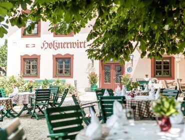 Außengastronomie Holznerwirt mit grünen Stühlen, karierten Tischdecken und einem Baum, der den Sitzbereich beschattet.