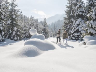 Zwei Menschen gehen durch tiefen Schnee in einem sonnenbeschienenen Wald in Altenmarkt-Zauchensee, umgeben von hohen Tannen.