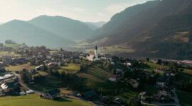 Luftaufnahme des kleinen Dorfes St.Veit im Pongau in der Region der Salzburger Sonnenterrasse mit einer Kirche und Bergen im Hintergrund bei Sonnenuntergang.