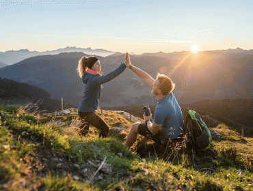 Zwei Wanderer geben sich bei Sonnenaufgang auf einem Berg die Hand, mit malerischen Tälern und fernen Gipfeln im Hintergrund.