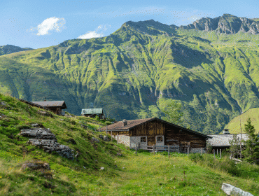 Holzhäuschen auf einem grasbewachsenen Hügel mit Bergen im Hintergrund unter blauem Himmel.