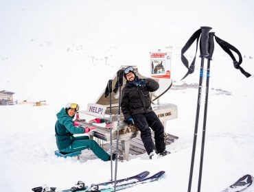 Zwei Skifahrer spielen auf einer verschneiten Piste auf einem Klavier mit der Aufschrift HELP! und lächeln dabei mit Skistöcken und Skiern in der Hand.