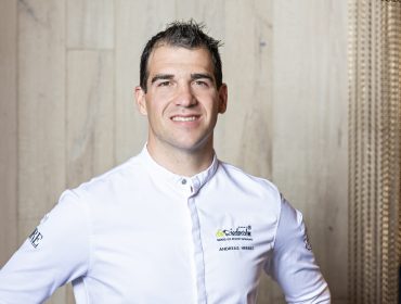 Eine lächelnde Köchin in weißer Uniform steht vor einer hellen, strukturierten Wand.