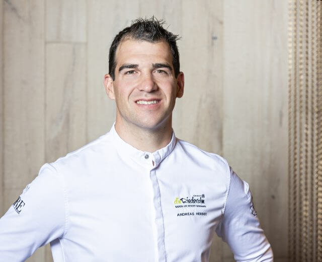 Eine lächelnde Köchin in weißer Uniform steht vor einer hellen, strukturierten Wand.