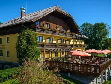 Ein gelbes Hotel mit Holzbalkonen und einer Terrasse mit roten Sonnenschirmen, umgeben von Grün an einem sonnigen Tag.
