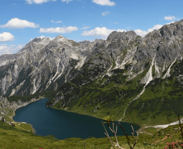 Ein tiefblauer Bergsee, umgeben von üppig grünen Hügeln und felsigen Gipfeln unter einem strahlend blauen Himmel.