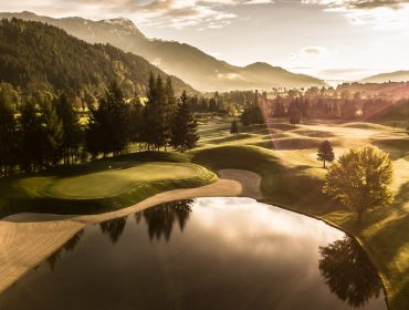 Ein Golfplatz bei Sonnenaufgang mit sanften Hügeln, Bäumen, einem Teich und Bergen im Hintergrund.