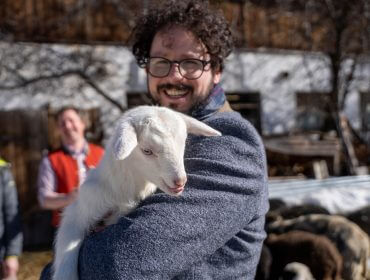 Rudi’s Roadtrip durch das SalzburgerLand – Winter Edition. Filzmoos Lamm Bratl, Gesenghofalm, Fiakerwirt Lächelnder Mann mit Brille hält eine weiße Babyziege an einem sonnigen Tag im Freien, mit Menschen und Tieren im Hintergrund.