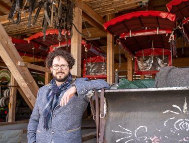 Ein Mann mit lockigem Haar und Brille lehnt sich an einen geschmückten Schlitten in einem rustikalen Holzstall mit rotgedeckten Kutschen.