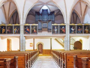 Innenraum einer Kirche mit Holzbänken, gewölbter Decke, Pfeifenorgel und gerahmten Porträts auf der Empore.