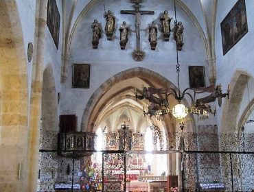 Gotisches Kircheninnere mit gewölbten Decken, Statuen, Altar und verzierten Metallarbeiten, die das Kirchenschiff vom Altarraum trennen.