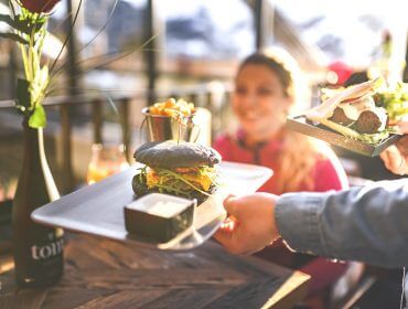 Ein Kellner bringt einer lächelnden Frau an einem sonnenbeschienenen Restauranttisch einen Burger mit schwarzem Brötchen und Pommes frites.