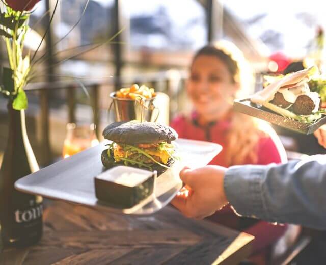 Ein Kellner bringt einer lächelnden Frau an einem sonnenbeschienenen Restauranttisch einen Burger mit schwarzem Brötchen und Pommes frites.