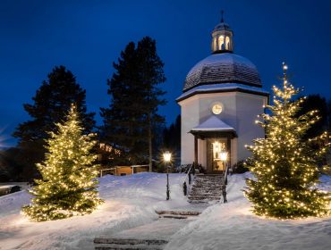 Eine verschneite Kapelle bei Nacht, mit Weihnachtsbäumen, die von Lichterketten auf beiden Seiten des Eingangs beleuchtet werden.