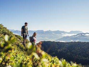 Zwei Personen wandern auf einem üppigen Bergpfad mit malerischem Blick auf ferne Berge unter einem klaren Himmel.