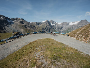 Zwei Radfahrer genießen das Gravel-Biken auf einer kurvigen Bergstraße mit schneebedeckten Gipfeln und klarem Himmel im Rücken.