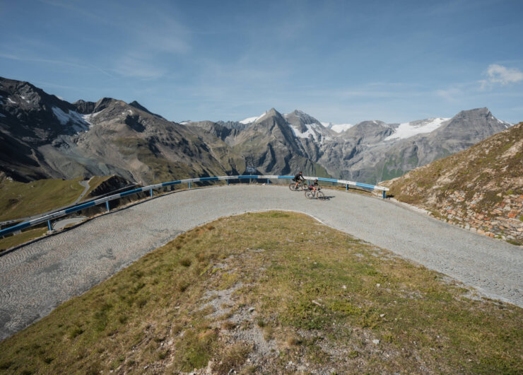 Zwei Radfahrer genießen das Gravel-Biken auf einer kurvigen Bergstraße mit schneebedeckten Gipfeln und klarem Himmel im Rücken.
