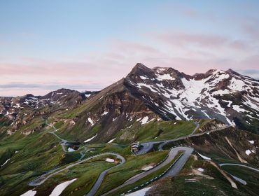 Eine kurvenreiche Bergstraße mit Schneeflecken, grünen Hügeln und einem pastellfarbenen Himmel bei Sonnenuntergang.