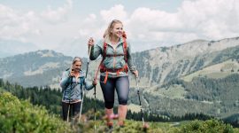 Zwei Frauen wandern mit Trekkingstöcken auf einem grünen Bergpfad mit malerischen Hügeln und Wolken im Hintergrund.
