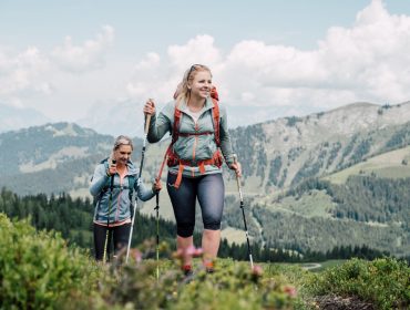 Zwei Frauen wandern mit Trekkingstöcken auf einem grünen Bergpfad mit malerischen Hügeln und Wolken im Hintergrund.