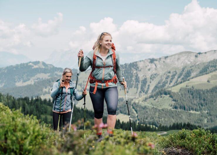 Zwei Frauen wandern mit Trekkingstöcken auf einem grünen Bergpfad mit malerischen Hügeln und Wolken im Hintergrund.