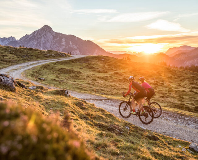 Zwei Gravel-Biker fahren bei Sonnenuntergang auf einem Bergweg, umgeben von grasbewachsenen Hügeln und fernen Gipfeln.