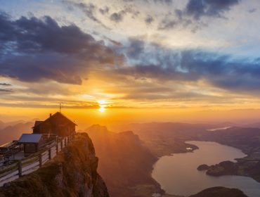 Sonnenuntergang über einer Berghütte am See, dramatische Wolken glühen, während ein Gravel-Biker durch die goldene Landschaft fährt.