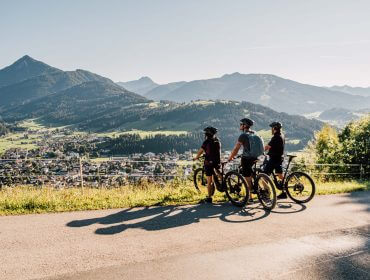 Drei Radfahrer machen auf einer Straße eine Pause, mit Blick auf eine malerische Berglandschaft und eine Talstadt im Sonnenlicht.