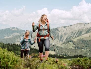 Zwei Frauen wandern mit Stöcken auf einem grünen Bergpfad, mit malerischen Hügeln und Wolken im Hintergrund.