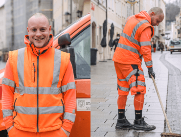 Ein lächelnder Straßenreiniger in einer orangefarbenen, reflektierenden Uniform steht da und fegt eine nasse Straße in der Stadt.