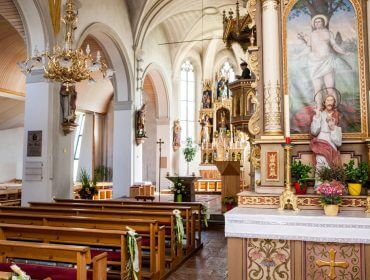 Innenraum einer geschmückten Kirche mit Holzbänken, Kronleuchtern und religiösen Gemälden auf dem Altar.